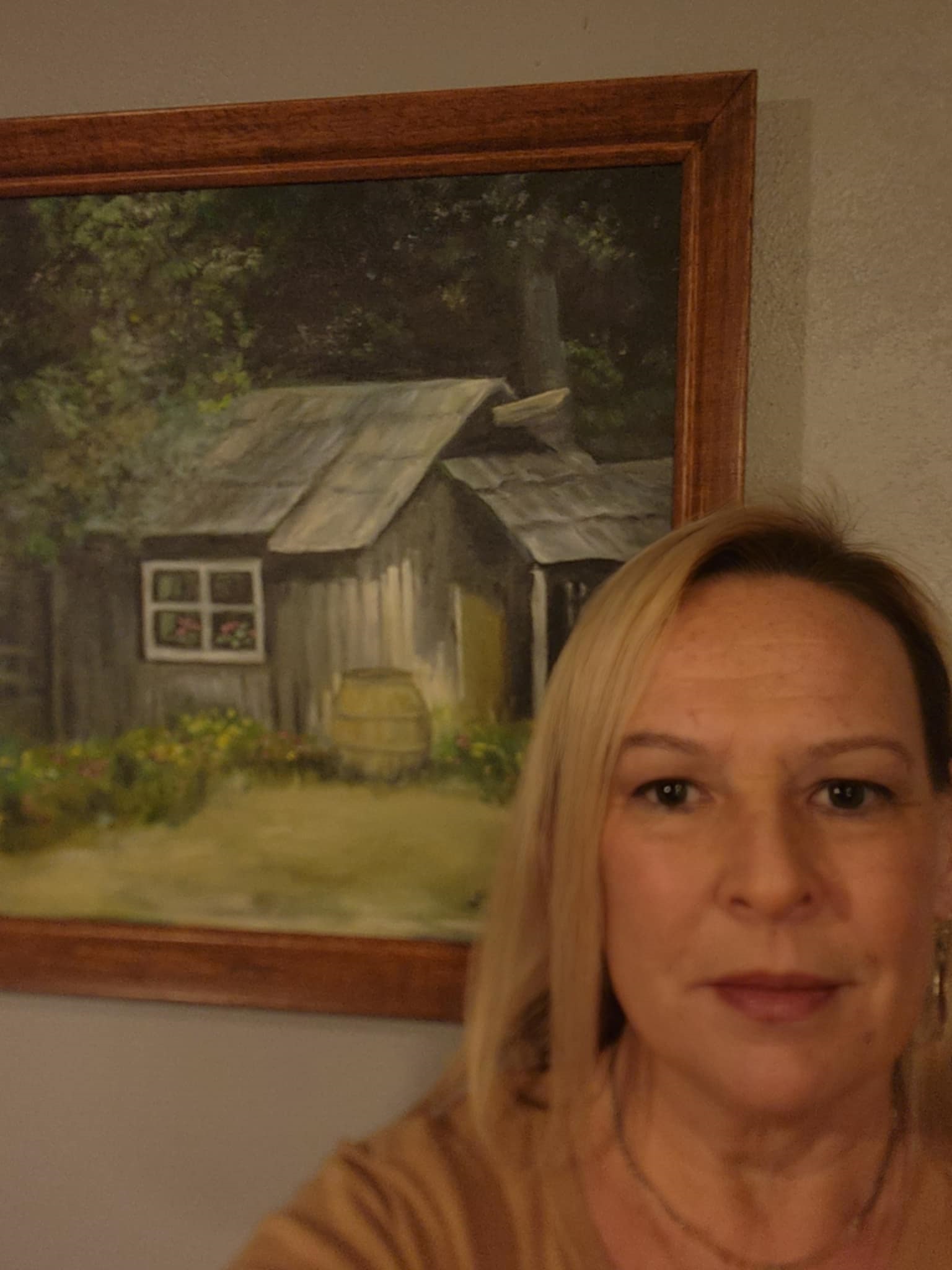 A woman with straight blonde hair stands in front of a framed painting of a rustic wooden cabin surrounded by trees and flowers.