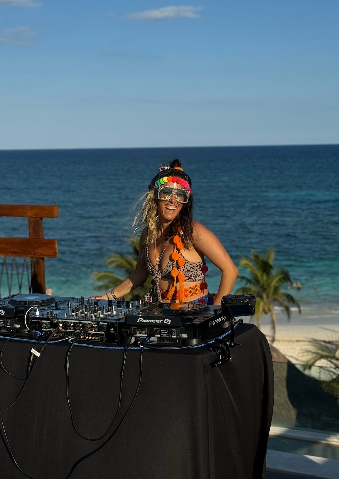 DJ Cheetah performing at an oceanfront venue in colorful festival attire with Pioneer DJ equipment, smiling against a tropical beach backdrop.