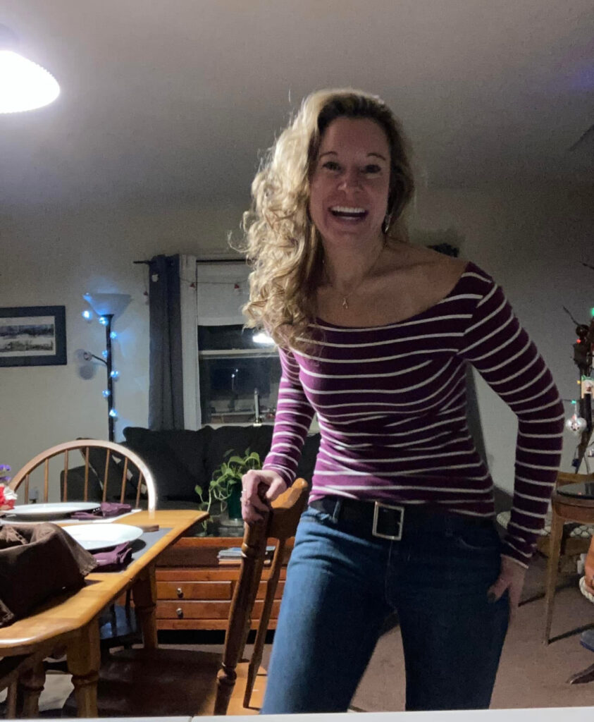 Woman with long curly hair wearing a maroon and white striped off-the-shoulder top and jeans, smiling while standing in a warmly lit living room.