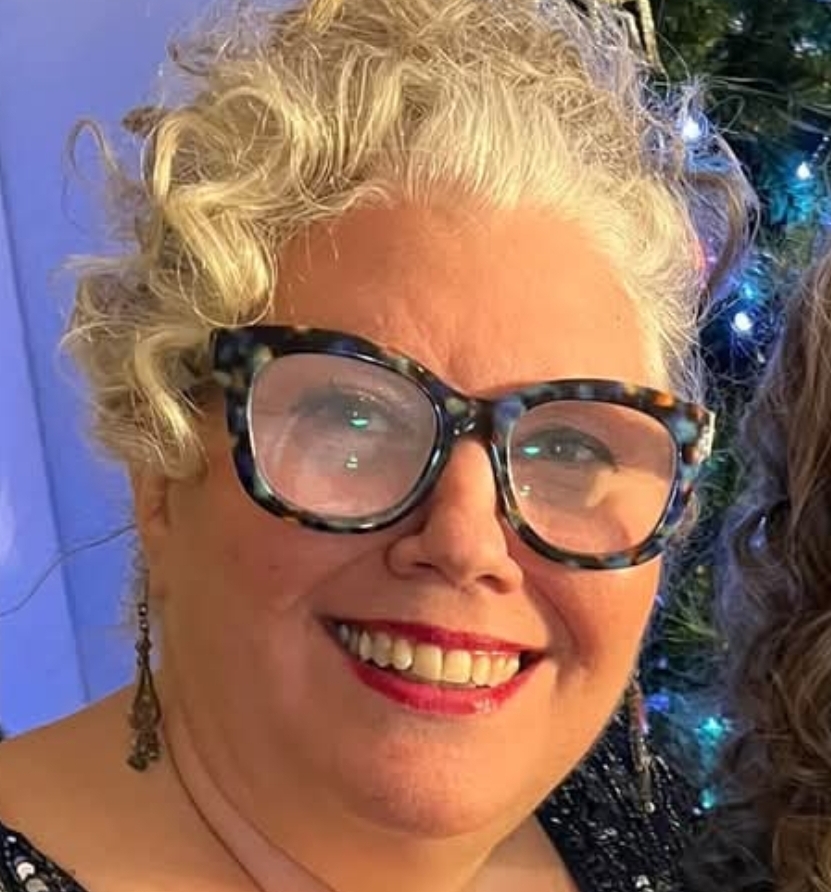 Cynthia smiling at a festive event, wearing bold glasses and red lipstick, with holiday lights in the background.