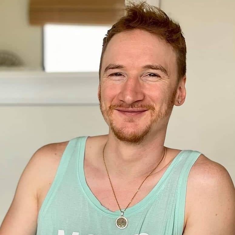 Smiling man with short red hair and a trimmed beard wearing a light turquoise tank top and a round pendant necklace, seated indoors in natural light
