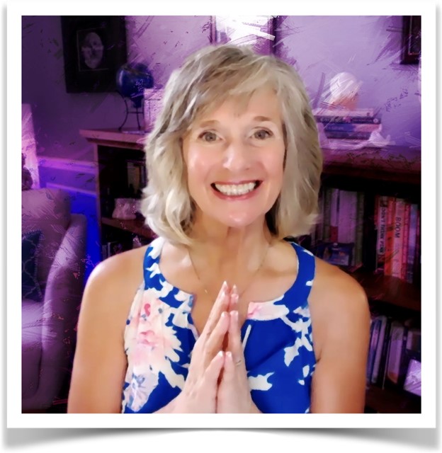 Smiling woman with short blonde hair wearing a blue floral dress, holding her hands in a prayer pose, standing in front of a bookshelf—Jeannie Lynch, Intuitive Energy Healer and Spirit Coach.
