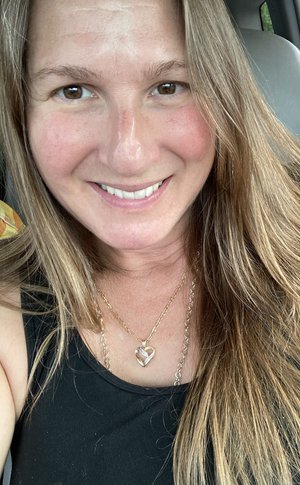 Smiling woman with long brown hair wearing a black tank top and a gold heart necklace, seated in a car with natural lighting.