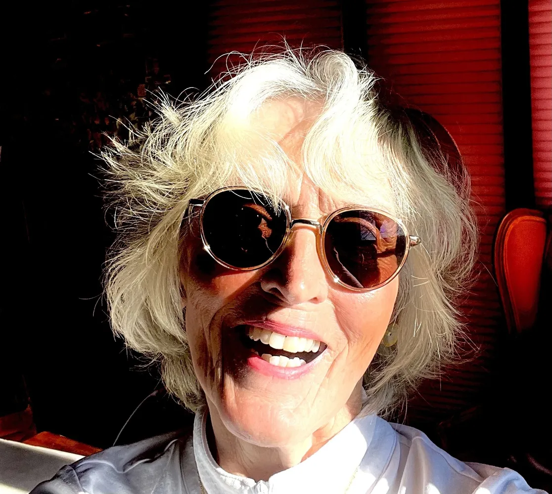 Smiling older woman with short, wavy white hair wearing round sunglasses and a white shirt, bathed in warm sunlight with a red-toned background.