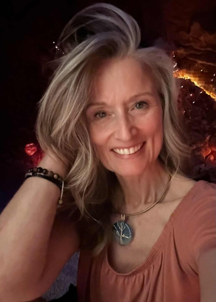 Smiling woman with long wavy blonde hair, wearing a peach-colored top and a pendant necklace featuring a tree design, posed in warm ambient lighting with twinkle lights in the background.