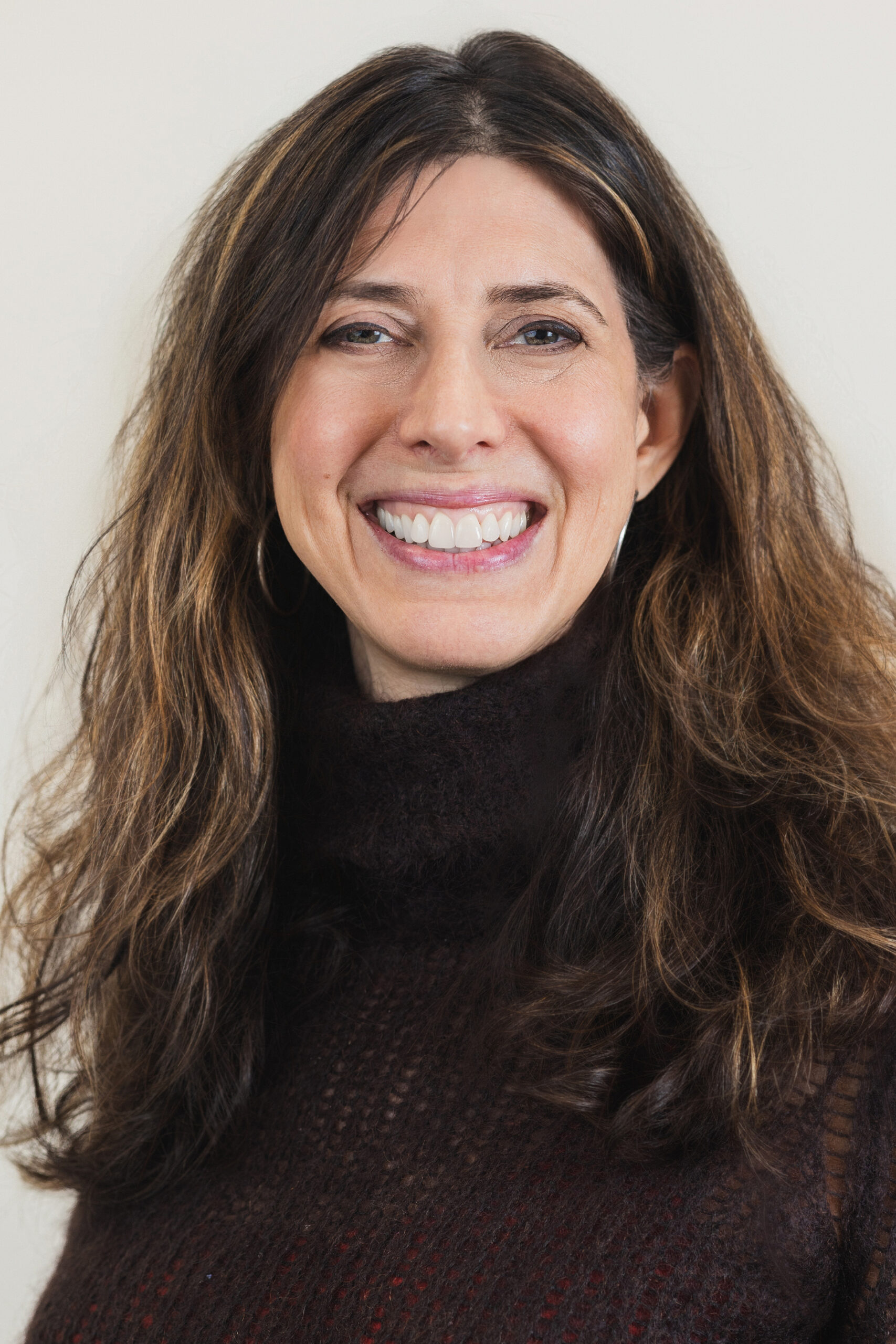 Professional headshot of a woman with long wavy brown hair, smiling warmly at the camera, wearing a dark textured sweater against a soft neutral background.