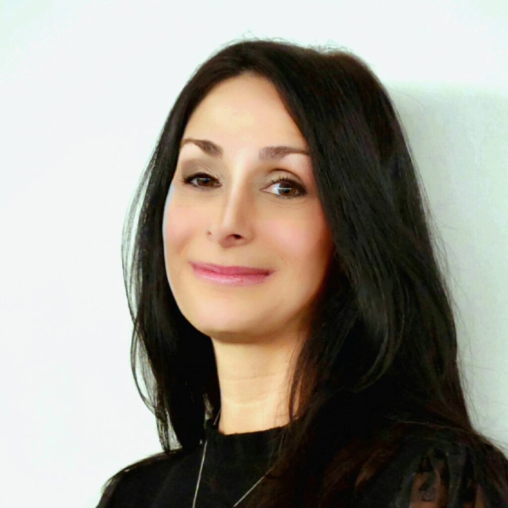 “Portrait of a woman with long dark hair, smiling softly, wearing a black top and a delicate necklace, against a light neutral background.”