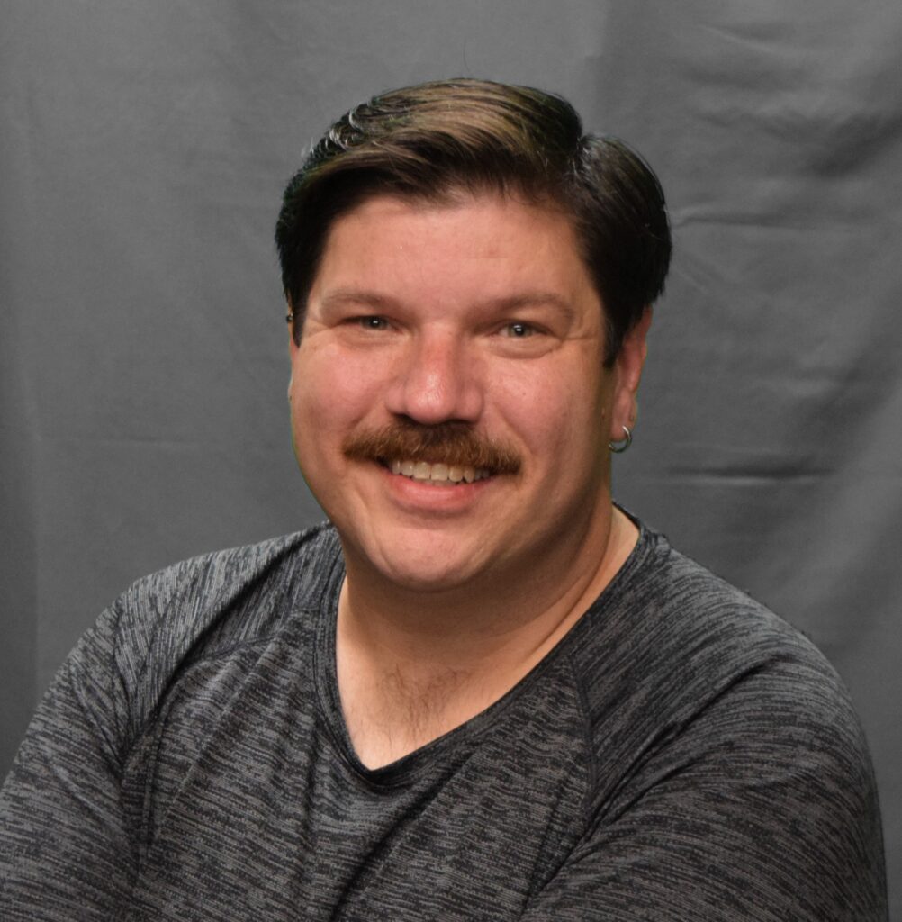 A man with short dark hair, a mustache, and light skin smiles warmly at the camera while wearing a dark gray textured shirt, set against a plain gray background.