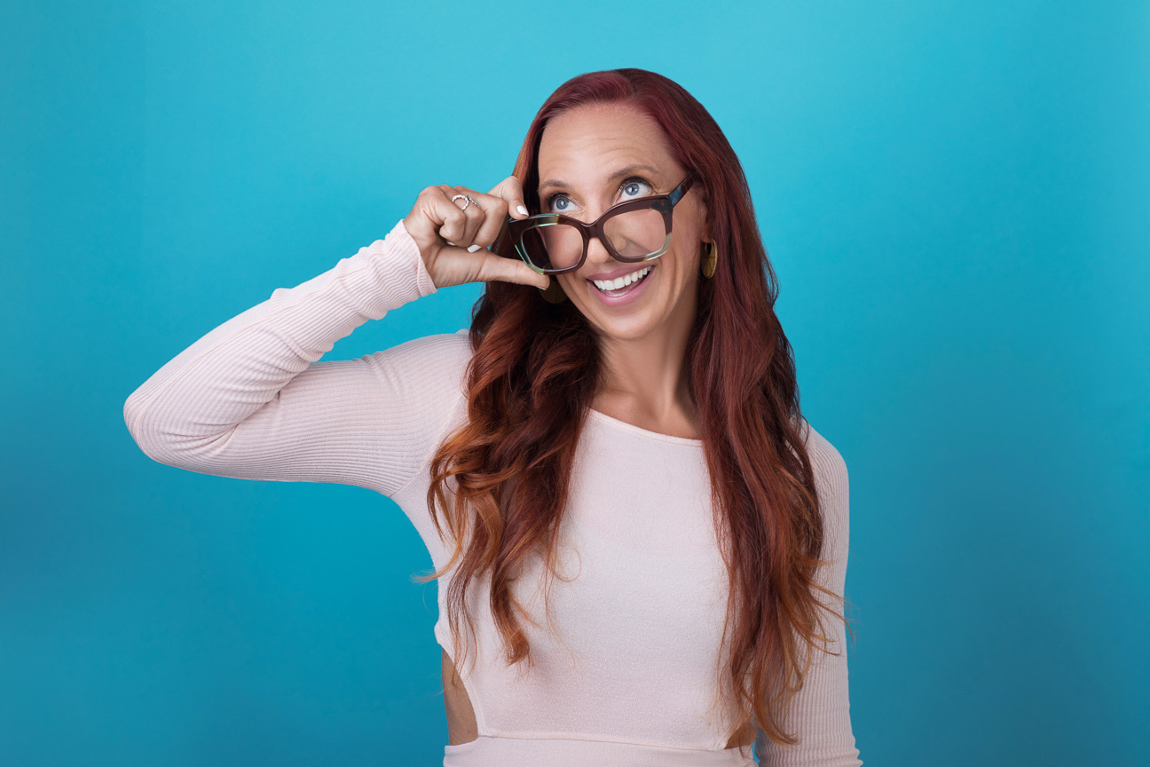 Long Red Haired woman in a pink shirt on a blue background with glasses, smiling