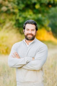 A bearded man with dark hair stands outdoors in soft golden-hour lighting, smiling with his arms crossed. He is wearing a light gray quarter-zip sweater, and the blurred background features trees and tall grass in warm green and yellow tones.