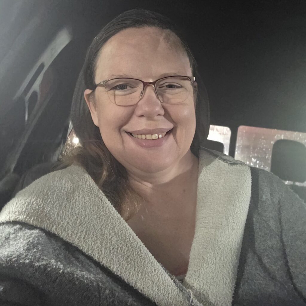 Long brown haired woman in a car smiling with a grey jacket on and thin glasses on her face
