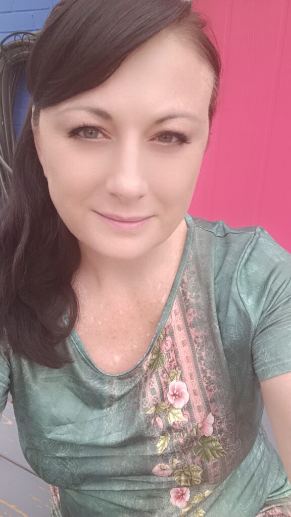 Long brown-haired light skinned woman taking a selfie with bright eyes and light smile on. she is wearing a green and pink tee shirt