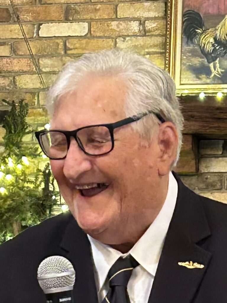 Elderly man in black suit with white shirt and grey hair and black glassed laughing behind a microphone