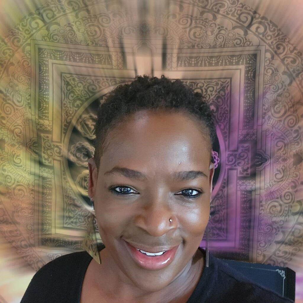 Beautiful short black-haired woman with warm brown skin, a big smile and a nose ring Infront of a wall tapestry. 
