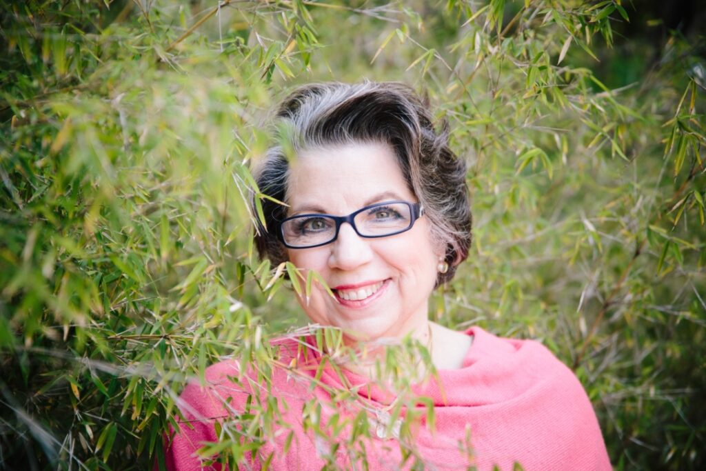 Middle aged light skinned woman wearing a pink shawl standing behind a willow tree. 